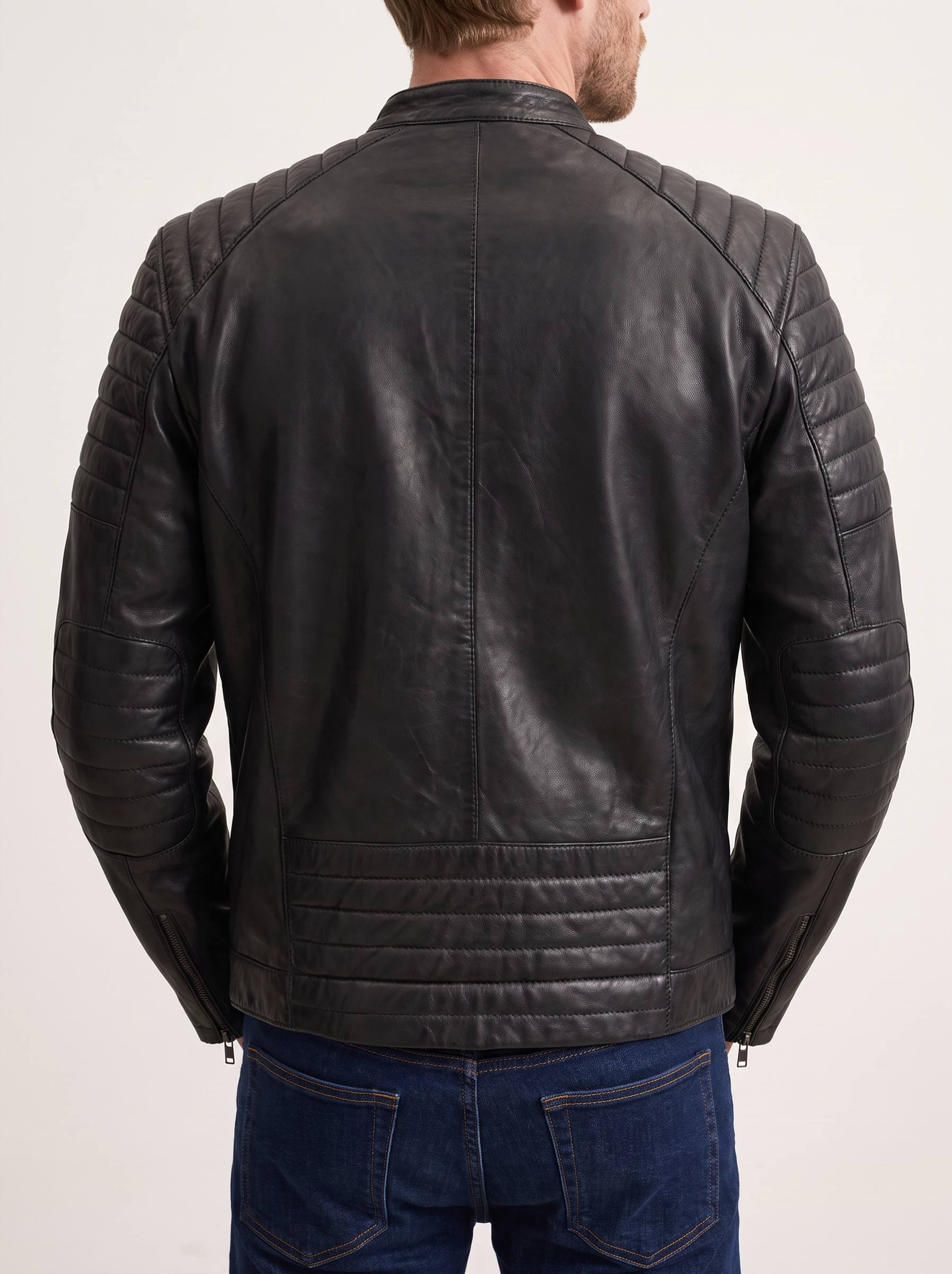 A man viewed from the back wearing a black leather jacket with quilted shoulder panels and horizontal stitching on the lower back, paired with blue jeans against a plain light background.