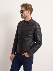 A man wearing a black leather motorcycle jacket with quilted shoulder details, paired with dark blue jeans and sunglasses, standing against a plain off-white background.