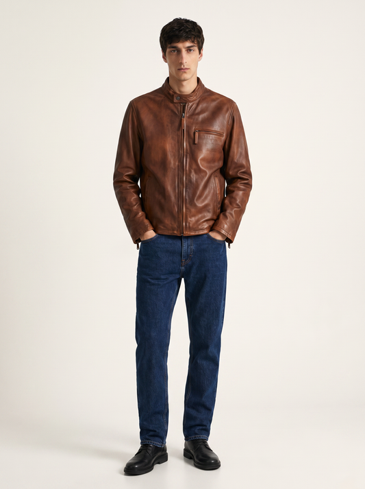 A man wearing a brown leather jacket, blue jeans, and black shoes, standing against a plain light background.