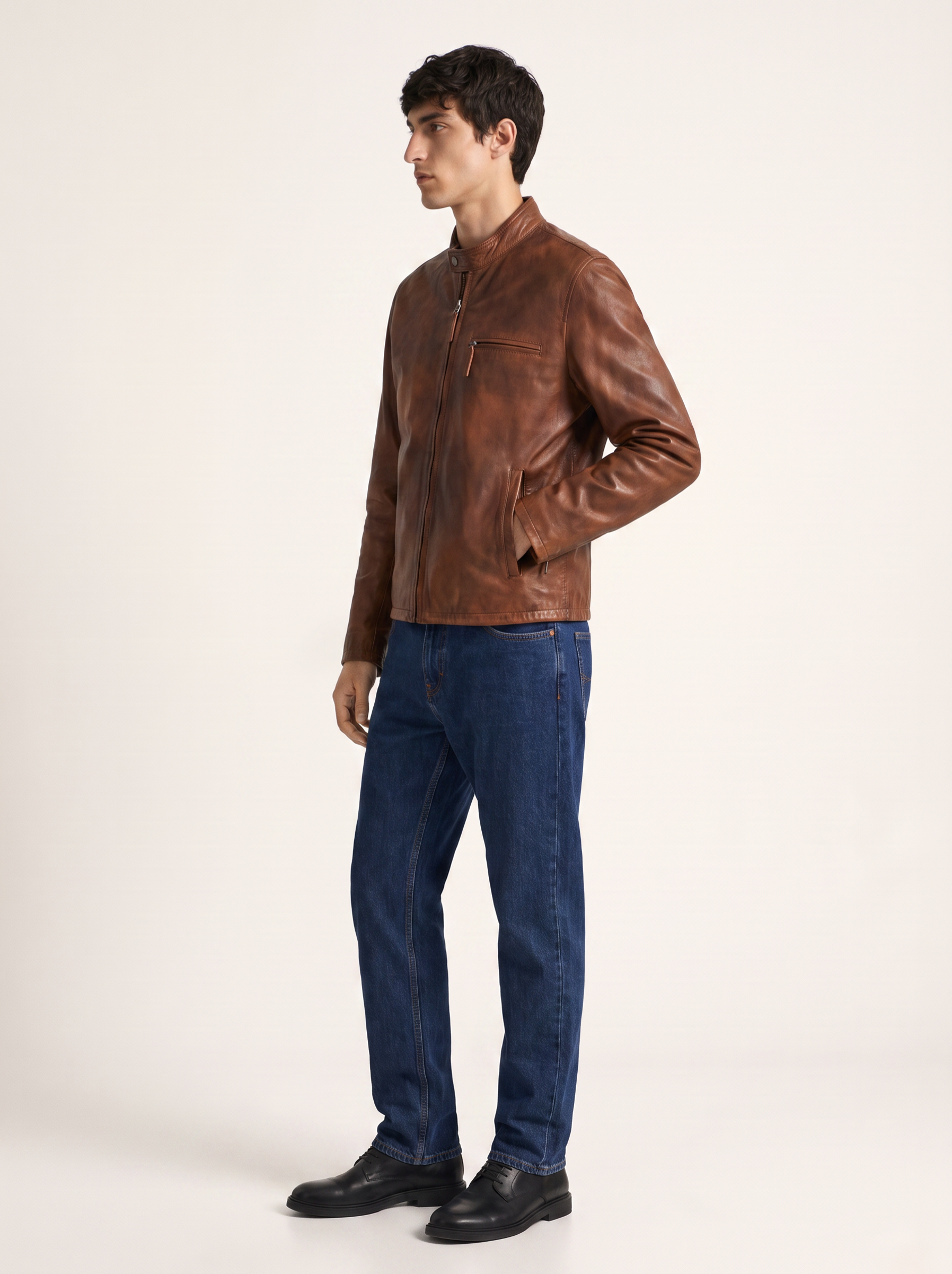 A man wearing a brown leather jacket, blue jeans, and black shoes, standing in profile against a plain off-white studio background.