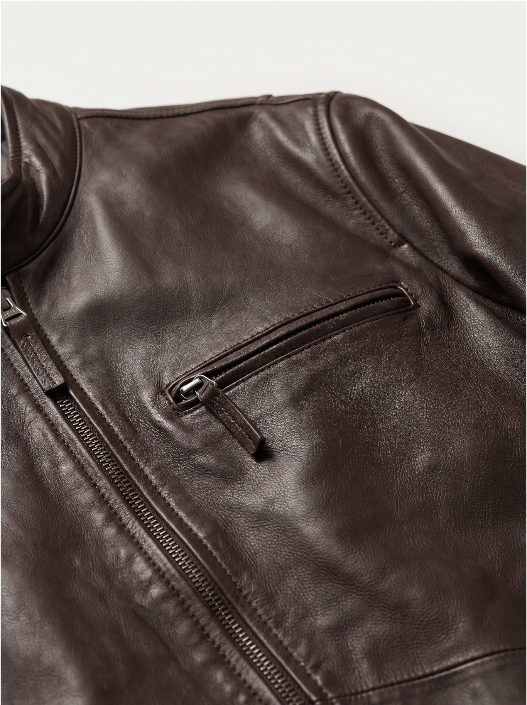 Close-up of a dark brown leather jacket showing a zippered pocket and part of the front zipper, photographed against a light neutral background.
