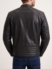A man viewed from the back wearing a black leather jacket with quilted shoulder panels and horizontal stitching on the lower back, paired with blue jeans against a plain light background.