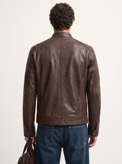 A man viewed from behind wearing a dark brown leather jacket and blue jeans, holding a brown leather bag against a plain off-white studio background.