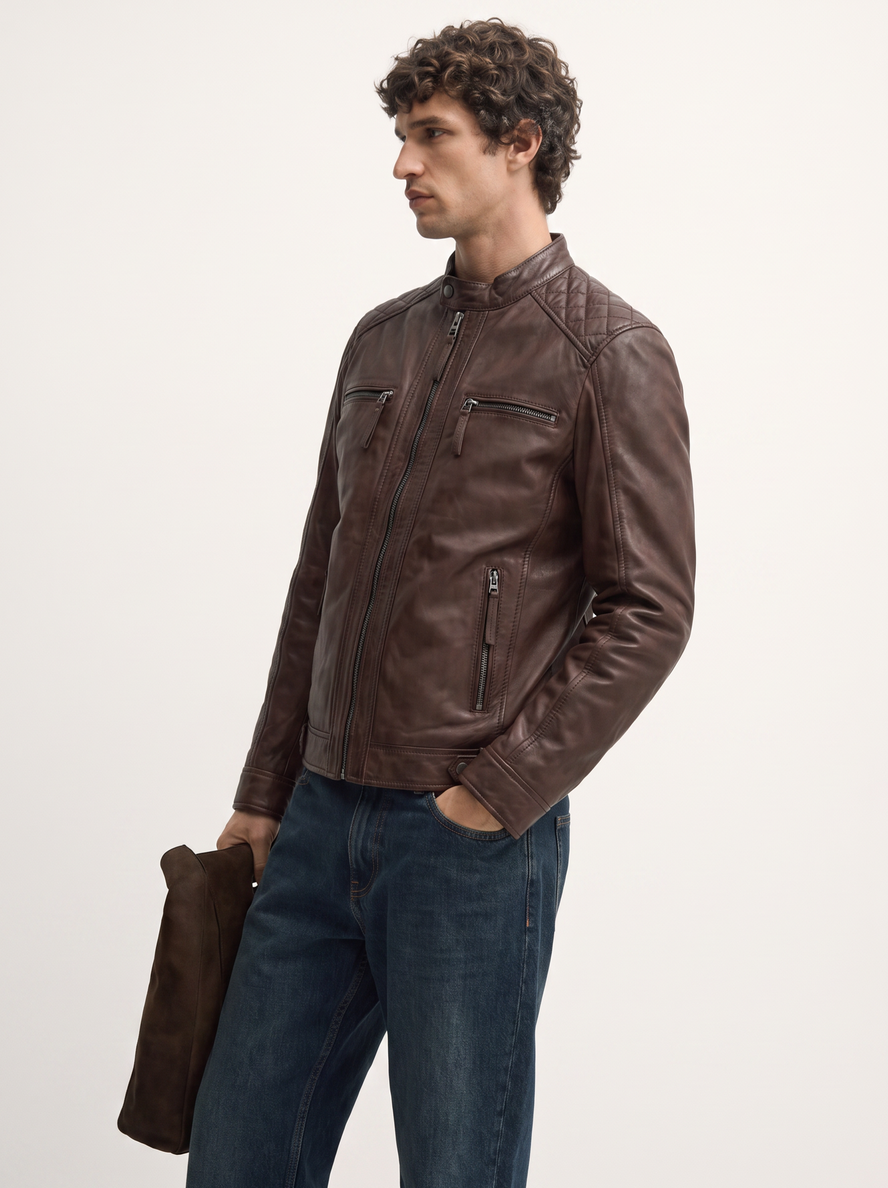A man wearing a dark brown leather jacket and blue jeans, holding a matching brown leather accessory against a plain white studio background.