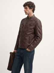 A man wearing a dark brown leather jacket and blue jeans, holding a matching brown leather accessory against a plain white studio background.