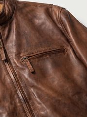 Close-up of a brown leather jacket showing the textured leather surface, a zippered front closure, and a horizontal zippered chest pocket.