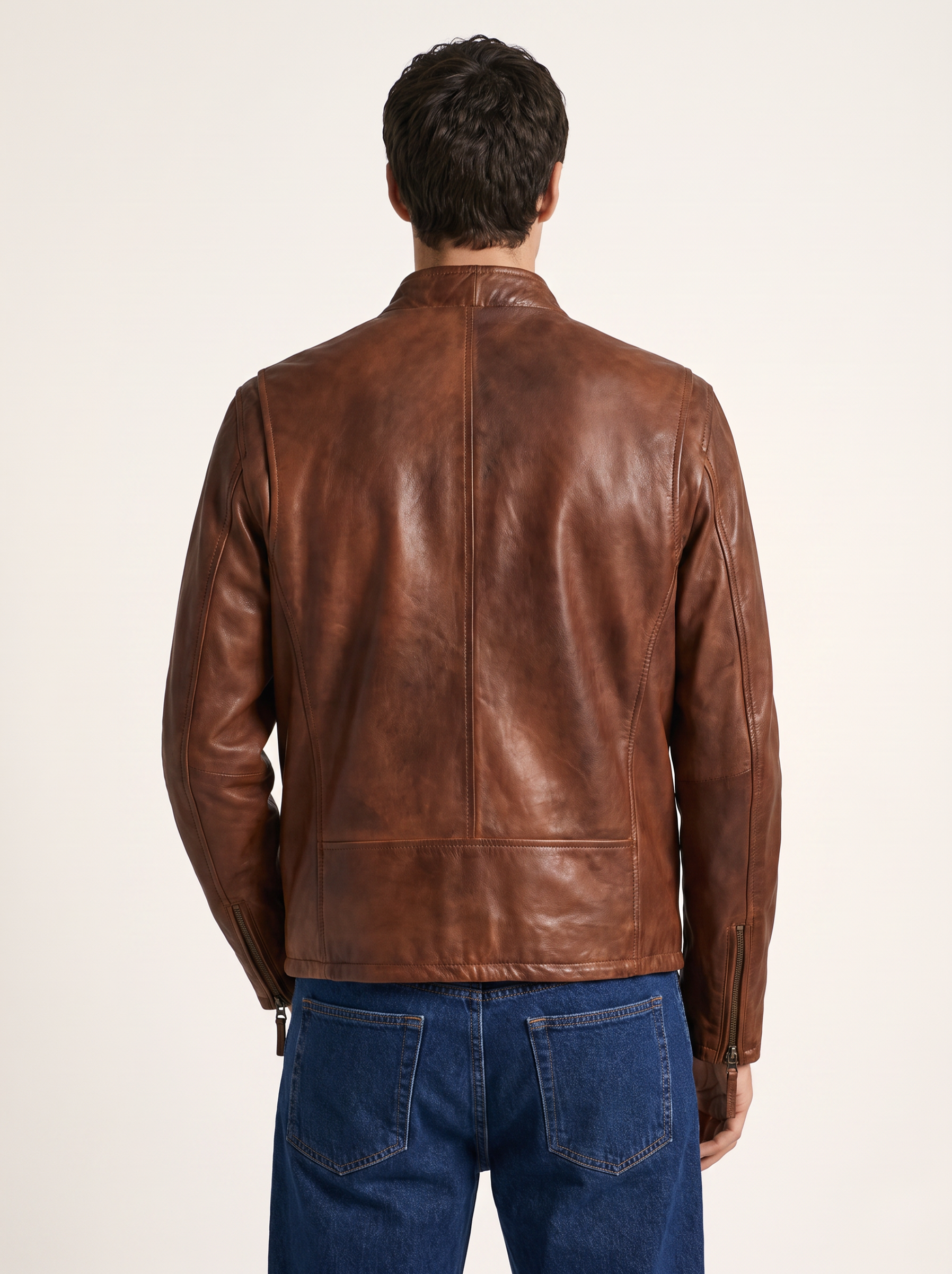 A man viewed from the back wearing a brown leather jacket and blue jeans, photographed against a plain light background.