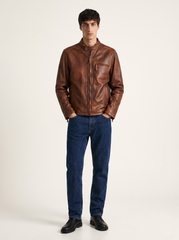A man wearing a brown leather jacket, blue jeans, and black shoes, standing against a plain light background.