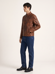 A man wearing a brown leather jacket, blue jeans, and black shoes, standing in profile against a plain off-white studio background.
