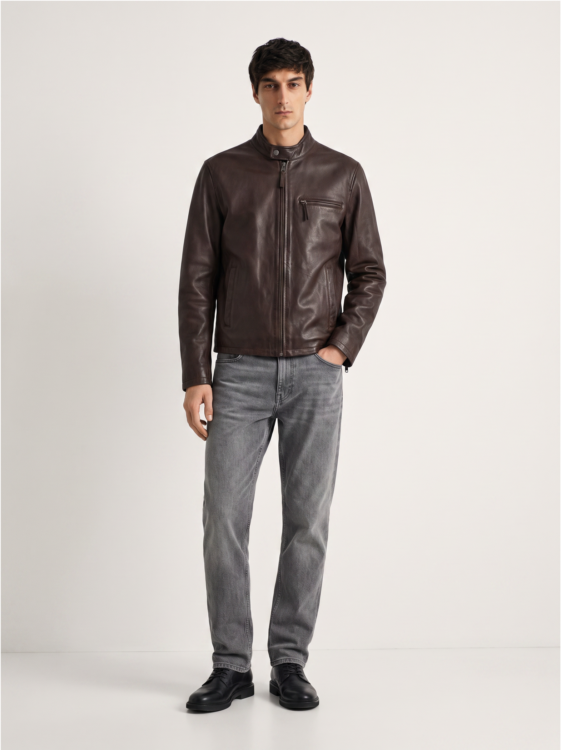 A man modeling a dark brown leather jacket with a stand collar and zippered chest pocket, paired with gray jeans and black shoes, standing against a light gray studio background.