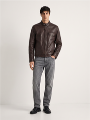 A man modeling a dark brown leather jacket with a stand collar and zippered chest pocket, paired with gray jeans and black shoes, standing against a light gray studio background.