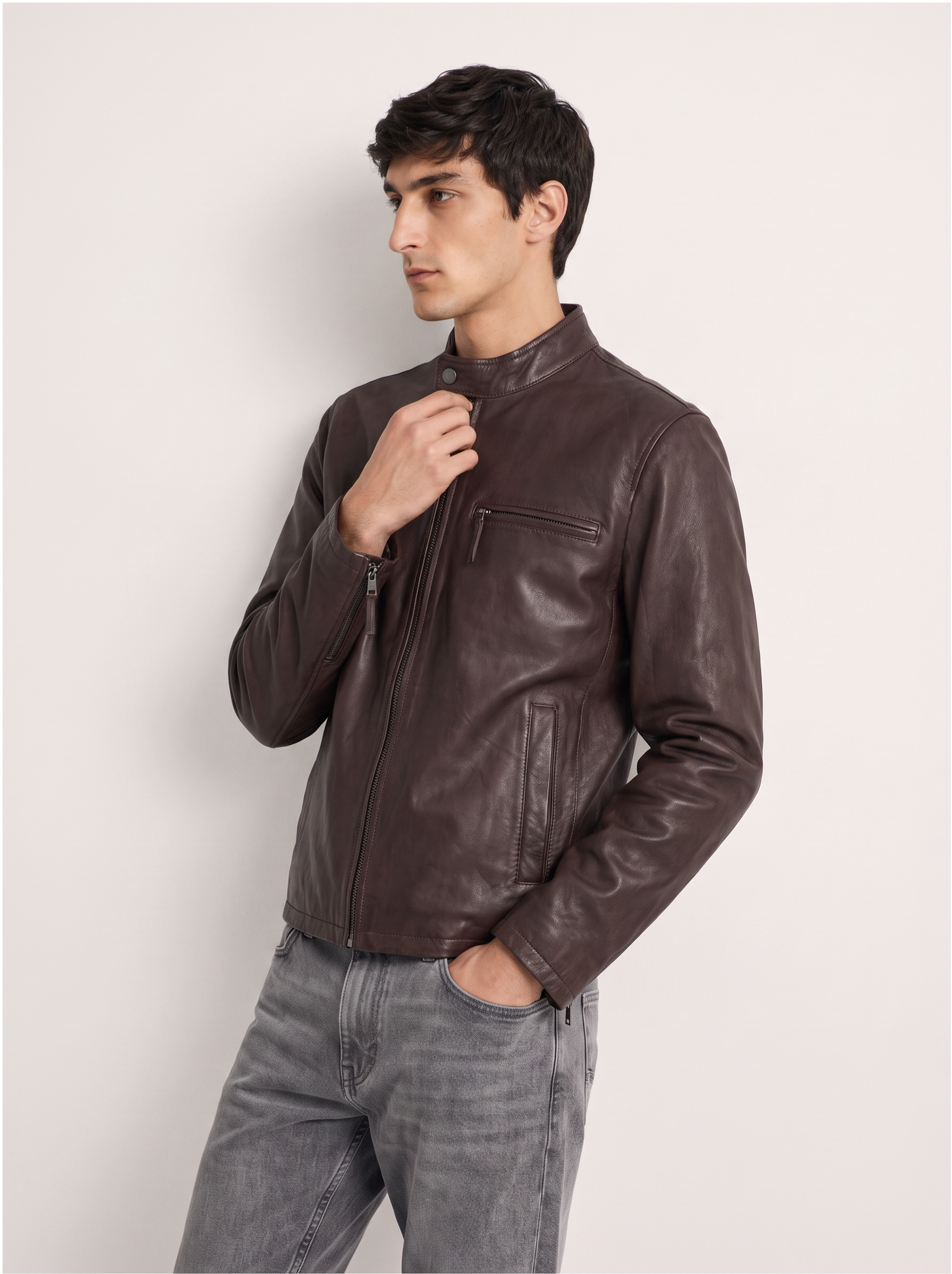 A man wearing a dark brown leather jacket and gray jeans, posing against a light off-white studio background.